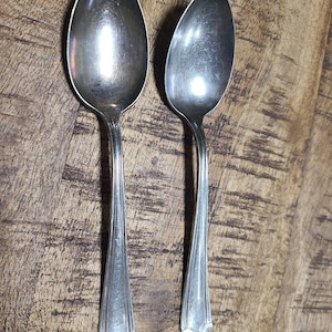 May include: Two silver spoons with ornate handles on a wooden surface.