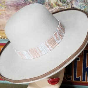 May include: A beige felt fedora hat with a brown leather band and a white and peach patterned band. The hat is sitting on a mannequin head.