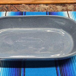 May include: A gray ceramic oval baking dish with handles. The dish has a smooth, glossy surface and is set on a blue and white striped surface. The dish is empty and ready for use.