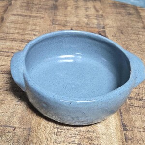 May include: A speckled, light blue ceramic bowl with two side handles. The bowl has a rounded shape and a smooth interior. The bowl is sitting on a wooden surface.