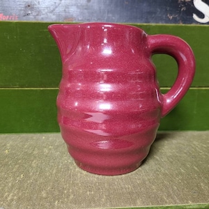 May include: A small, red, ceramic pitcher with a ribbed design. The pitcher has a handle and a spout.