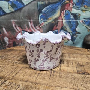 May include: A white ceramic planter with a ruffled rim, adorned with a speckled purple pattern. The planter sits on a wooden surface, with a backdrop featuring a mural of blue fish.