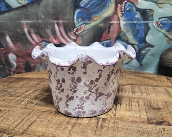 May include: A white ceramic planter with a ruffled rim, adorned with a speckled purple pattern. The planter sits on a wooden surface, with a backdrop featuring a mural of blue fish.
