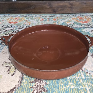 May include: A round, brown metal tray with two handles. The tray has a shallow depth and a smooth surface. The handles are small and curved. The tray is sitting on a colorful patterned rug.