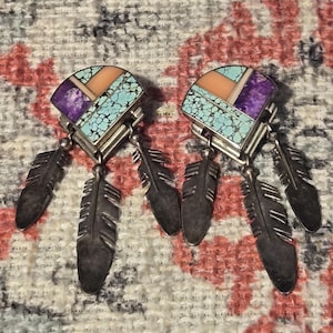 May include: A pair of silver earrings with a turquoise, purple, and peach-colored mosaic design. Each earring has three dangling silver feathers. The earrings are displayed on a patterned fabric background.