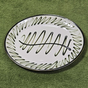 May include: White oval ceramic plate with a black and green fern design. The plate has a black rim with a gold accent.