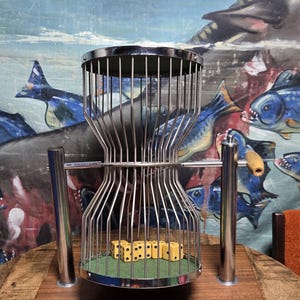 May include: A chrome-plated metal dice cage with vertical bars, mounted on a rectangular wooden base. The cage features a rotating mechanism with a wooden handle and contains several yellow six-sided dice resting on a green felt floor.