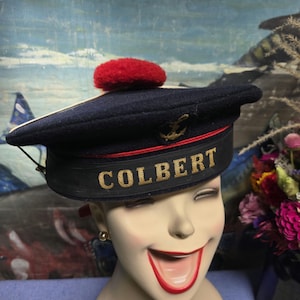 May include: A navy blue sailor hat with a red pom-pom on top. The hat has a gold anchor and the word "COLBERT" embroidered on the front.