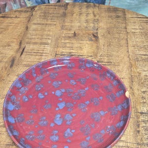 May include: A round ceramic plate with a maroon base and blue speckled design. The plate is on a wooden surface, with a painting of blue fish in the background.