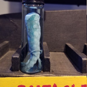 May include: A clear glass jar with a black lid containing a preserved tentacle in a blue liquid. The jar is labeled "TENTACLE".