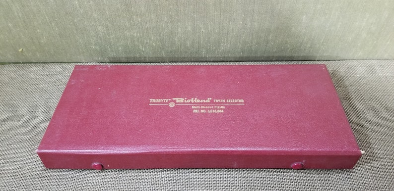 May include: A burgundy rectangular box with the text "TRUBYTE Bioland TRY-ON SELECTOR" in gold lettering. The box is closed and has a textured surface. The box also has the text "Multi-Shade Plastic" and "PAT. NO. 3,015,884".