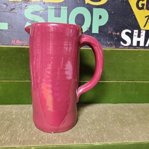 May include: A deep red ceramic pitcher with a handle. The pitcher is made of a smooth, glossy material.