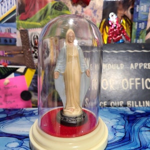 May include: A religious figurine of a woman with outstretched arms, encased in a clear glass dome. The figurine is beige with light blue accents, set on a red base and a cream-colored, tiered stand. The background includes art and a red bow.