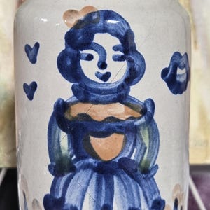 May include: A ceramic jar with a hand-painted design of a woman in a blue dress. The jar has a white base with blue and orange accents. The background features a painting of an angel.
