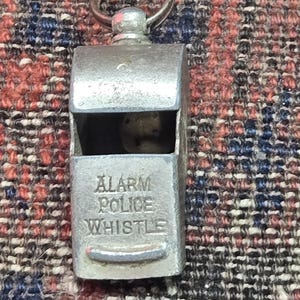 May include: A vintage silver-toned whistle with a ring for attachment. The text "ALARM POLICE WHISTLE" is embossed on the front. The whistle is rectangular with a small opening at the top.