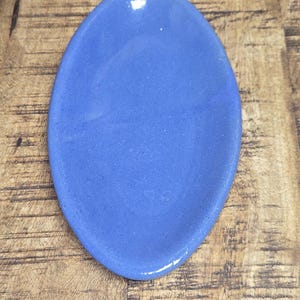 May include: A small, oval-shaped, ceramic dish in a vibrant blue hue. The dish has a smooth, glossy finish and is likely used for holding small items. The background is a wooden surface.