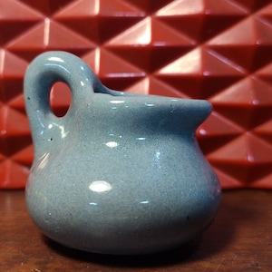 May include: A small, light blue ceramic creamer with a handle and spout. The creamer has a round body and a speckled texture. The handle is a loop shape. The background features a red geometric design.