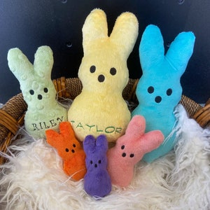 Pode incluir: Cinco brinquedos de pelúcia em forma de coelho em uma cesta de vime forrada com pele sintética branca. Os coelhos são de várias cores, incluindo amarelo, verde, azul, laranja e roxo. O coelho amarelo tem o nome "TAYLOR" bordado e o coelho verde tem o nome "RILEY" bordado.