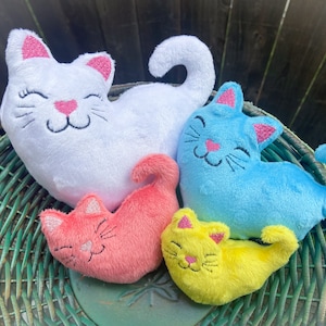 以下が含まれることがあります： 籐のバスケットの中に、ぬいぐるみ製の猫の玩具が4つあります。猫は白、青、ピンク、黄色です。それぞれの猫は刺繍された目と笑顔の口をしています。