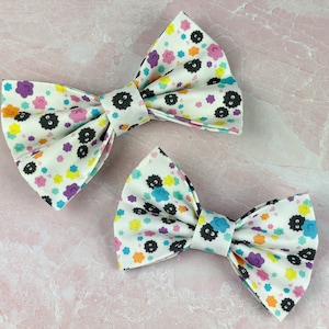 May include: Two white fabric bow ties with a black and colorful floral pattern. The pattern features small black cartoon faces with white eyes.