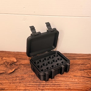 May include: A matte black rectangular storage case with a hinged lid and two front latches. The interior base features a grid of fifteen circular recessed compartments designed for holding small items, with a smooth, flat underside on the lid.