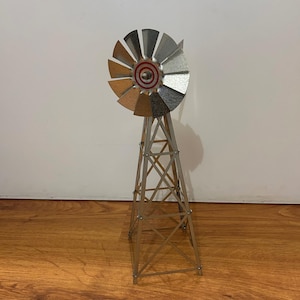 May include: A metallic windmill model featuring a tall, four-legged lattice tower structure supporting a circular fan with multiple silver blades. The center of the fan displays a small, circular red and white target-like graphic design.
