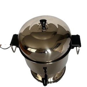 May include: A polished stainless steel coffee percolator with a rounded lid, a black knob handle on top, and two black side handles. The cylindrical body features a black spigot at the base for dispensing liquid, reflecting the surrounding environment.