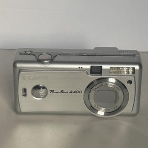 May include: A silver Canon PowerShot A400 digital camera featuring a 3.2 megapixel lens, a built-in flash, and a zoom lens with 5.9-13.2mm 1:3.8 specifications. The top panel includes a power button and a shutter release button.