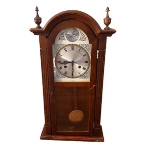 May include: A brown wooden mantel clock with a silver-toned circular face featuring Roman numerals and the text Tempus Fugit. The clock has a glass-fronted lower section revealing a pendulum and two decorative finials on the arched top.