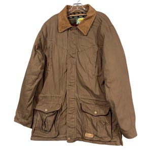 May include: A brown canvas field jacket featuring a tan corduroy collar, a front button placket, and two large lower cargo pockets with buttoned flaps. The jacket includes a Cabela's brand label on the interior neck and a small logo patch on the lower right pocket.