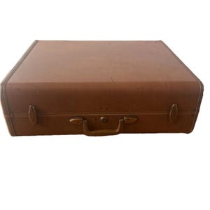 Pode incluir: Uma mala retangular de cor castanha clara com um acabamento exterior liso. A bagagem possui uma pega central, dois fechos laterais e um mecanismo de bloqueio central com fechadura. As bordas são reforçadas com um rebordo a condizer.
