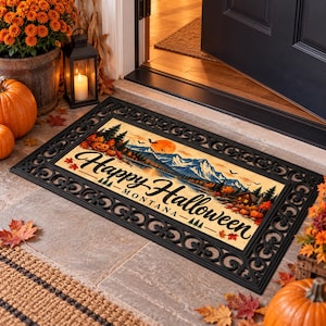 Puede incluir: Un felpudo rectangular con un borde decorativo negro y un gráfico central que muestra un paisaje montañoso, una luna llena, calabazas y árboles. El texto dice Happy Halloween MONTANA en letras negras.