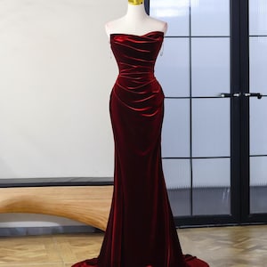 May include: A floor-length, strapless gown in deep red velvet. The bodice features a draped, cowl-style neckline with intricate ruching that extends down the torso, creating a fitted silhouette that flares slightly at the hem into a small train.