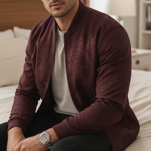 May include: A deep burgundy bomber jacket with a subtle tonal paisley pattern, featuring a ribbed collar and cuffs, worn over a light grey crew neck t-shirt, paired with black trousers and a silver-toned metal link wristwatch on the left wrist.