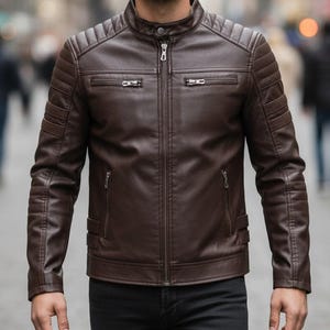 May include: A dark brown leather jacket featuring a stand-up collar with a snap button, a central zipper closure, and two horizontal zippered chest pockets. The shoulders and upper sleeves have quilted padding, and there are two lower pockets.