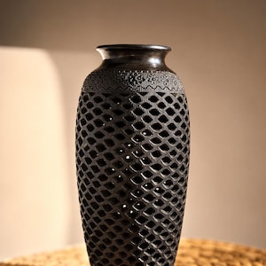 Puede incluir: Un jarrón negro de forma cilíndrica con un borde ensanchado. El cuerpo presenta un intrincado patrón de celosía calada en el centro, mientras que la sección superior está decorada con una banda horizontal de pequeños motivos florales tallados.