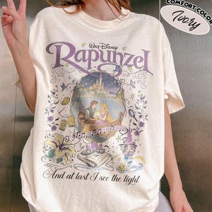 May include: An ivory t-shirt featuring a graphic print of the Disney movie Rapunzel. The design includes the title Rapunzel, a scene of two characters in a boat, a castle, flowers, a book, and the text And at last I see the light.