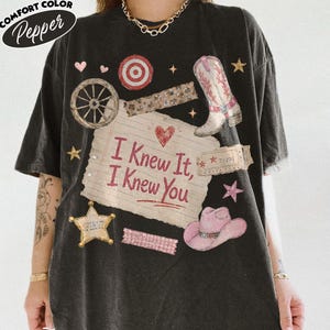 Op de afbeelding: Een zwart t-shirt met korte mouwen en een centrale afbeelding van een gelinieerd notitieblaadje met de tekst "I Knew It, I Knew You", omringd door illustraties van een cowboylaars, een roze cowboyhoed, een wagenwiel en kleine hartjes.