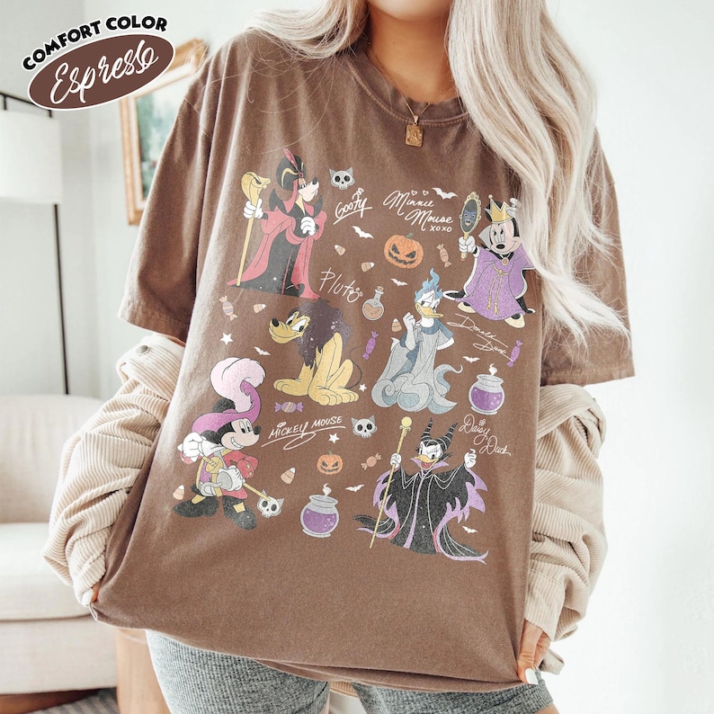 May include: An espresso brown short-sleeve t-shirt featuring a graphic print of various Disney characters in Halloween costumes, including Mickey Mouse, Minnie Mouse, Donald Duck, Goofy, Pluto, and Maleficent, along with pumpkins and candy icons.