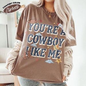 Peut inclure: Un t-shirt marron &agrave; manches courtes avec le texte "YOU'RE A COWBOY LIKE ME" en lettres bleues. Le motif comprend des illustrations d'un chapeau de cowboy, d'une fus&eacute;e, d'une balle, de bottes, de nuages et d'une &eacute;toile de sh&eacute;rif en jaune et rouge.