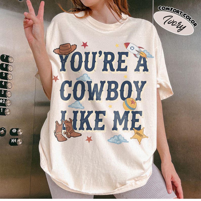 Peut inclure: Un t-shirt &agrave; manches courtes ivoire avec le texte bleu fonc&eacute; "YOU'RE A COWBOY LIKE ME" entour&eacute; d'illustrations graphiques d'un chapeau de cowboy marron, d'une fus&eacute;e, d'une balle color&eacute;e, d'une &eacute;toile de sh&eacute;rif, de petites &eacute;toiles rouges et de nuages bleu clair.