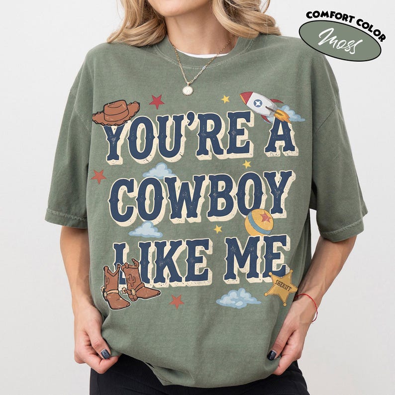 Peut inclure: Un t-shirt &agrave; manches courtes vert mousse avec le texte "YOU'RE A COWBOY LIKE ME" en lettres bleu fonc&eacute;. Le motif comprend des illustrations d'un chapeau de cowboy marron, d'une fus&eacute;e, d'une balle, de bottes, d'une &eacute;toile de sh&eacute;rif, de nuages et d'&eacute;toiles.