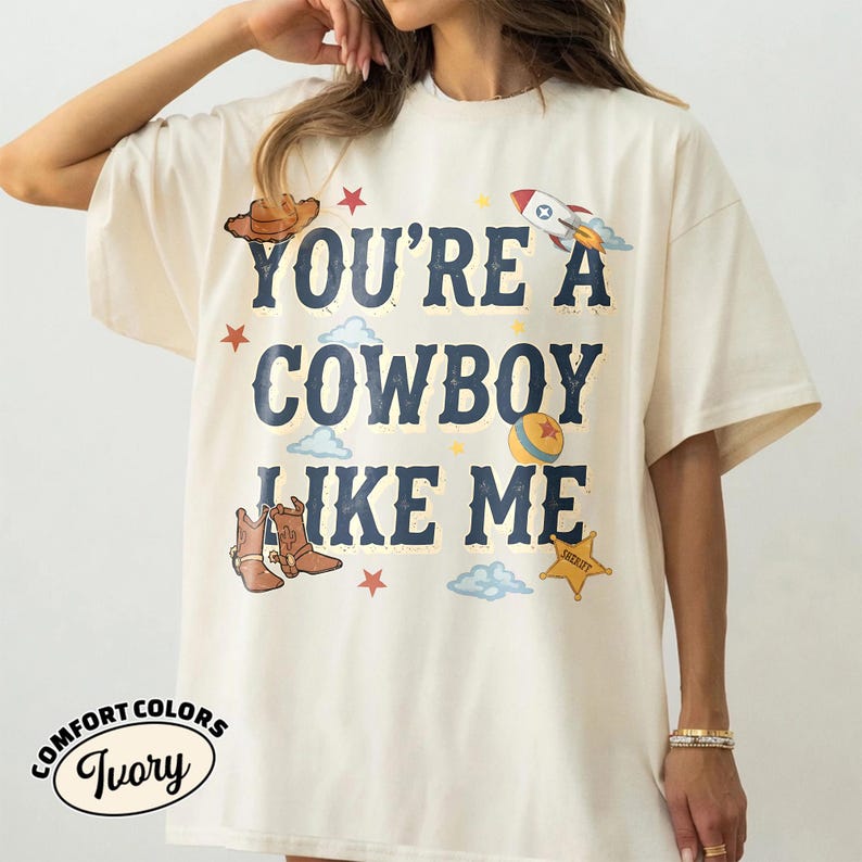 Peut inclure: Un t-shirt ivoire avec le texte bleu fonc&eacute; YOU'RE A COWBOY LIKE ME. Le motif comprend un chapeau de cowboy marron, une fus&eacute;e, une balle, une &eacute;toile de sh&eacute;rif, ainsi que des petits nuages et des &eacute;toiles dispers&eacute;s autour du lettrage.