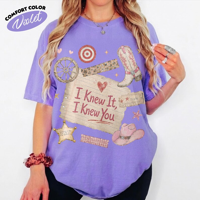 Op de afbeelding: Een violetkleurig T-shirt met korte mouwen en een grafische print van een gelinieerd briefje met de tekst "I Knew It, I Knew You" en een rood hart. Het ontwerp bevat een cowboylaars, een hoed, een wagenwiel, een ster en een doelwit.