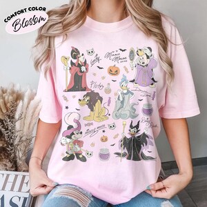 May include: A light pink t-shirt featuring a graphic print of various cartoon characters dressed in Halloween costumes. The design includes illustrations of characters, pumpkins, candies, and skulls, along with handwritten-style names and text.