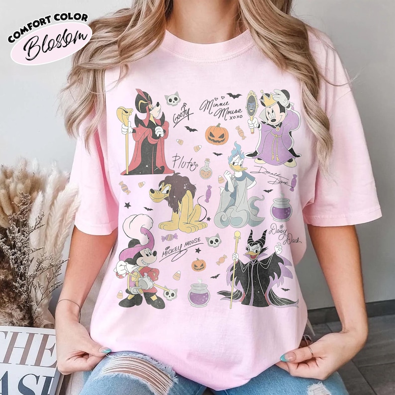 May include: A light pink t-shirt featuring a graphic print of various cartoon characters dressed in Halloween costumes. The design includes illustrations of characters, pumpkins, candies, and skulls, along with handwritten-style names and text.