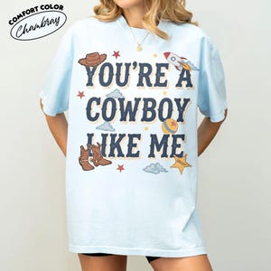 Peut inclure: Un t-shirt bleu clair avec le texte bleu fonc&eacute; YOU'RE A COWBOY LIKE ME. Le motif comprend un chapeau de cowboy marron, une fus&eacute;e, une balle, une &eacute;toile de sh&eacute;rif et des nuages. Le t-shirt a des manches courtes et un col rond.