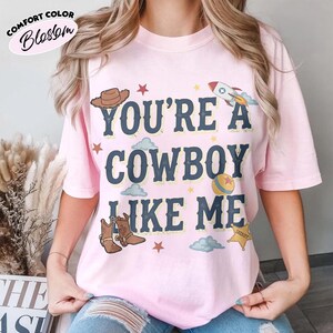 Peut inclure: Un t-shirt rose clair &agrave; manches courtes avec le texte bleu fonc&eacute; "YOU'RE A COWBOY LIKE ME" entour&eacute; d'illustrations graphiques d'un chapeau de cowboy marron, d'une fus&eacute;e, d'une balle jaune, d'une &eacute;toile de sh&eacute;rif, d'&eacute;toiles rouges et de nuages bleu clair.