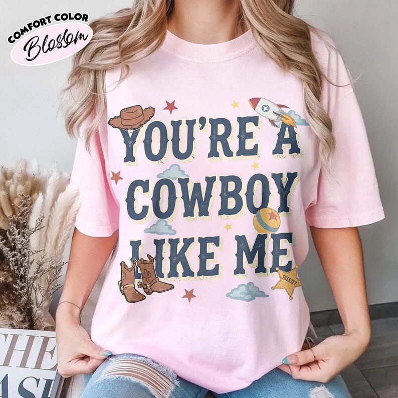 Peut inclure: Un t-shirt rose clair &agrave; manches courtes avec le texte bleu fonc&eacute; "YOU'RE A COWBOY LIKE ME" entour&eacute; d'illustrations graphiques d'un chapeau de cowboy marron, d'une fus&eacute;e, d'une balle jaune, d'une &eacute;toile de sh&eacute;rif, d'&eacute;toiles rouges et de nuages bleu clair.