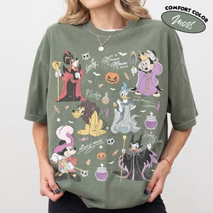May include: A moss green short-sleeve t-shirt featuring a graphic print of various Disney characters in Halloween costumes, including Mickey Mouse, Minnie Mouse, Donald Duck, and Pluto, alongside pumpkins, candy, and text like Minnie Mouse XOXO.